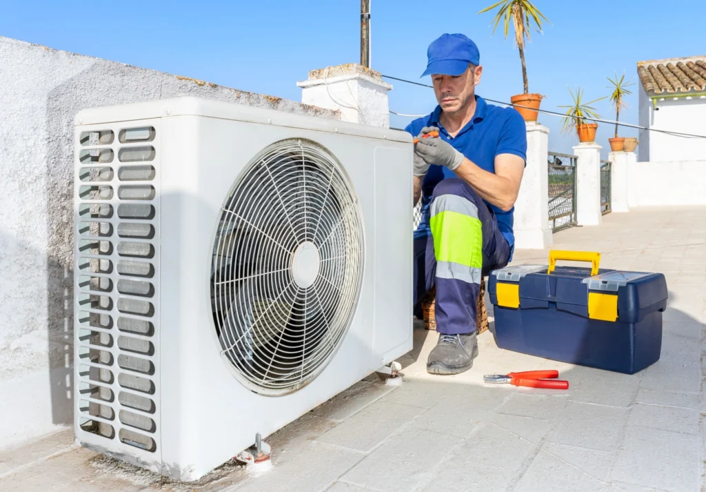 An HVAC technician from Family Heating, Cooling, Electric & Plumbing in a blue uniform and cap using hand tools to service a white outdoor air conditioning condenser unit on a sunny rooftop next to his blue toolbox.