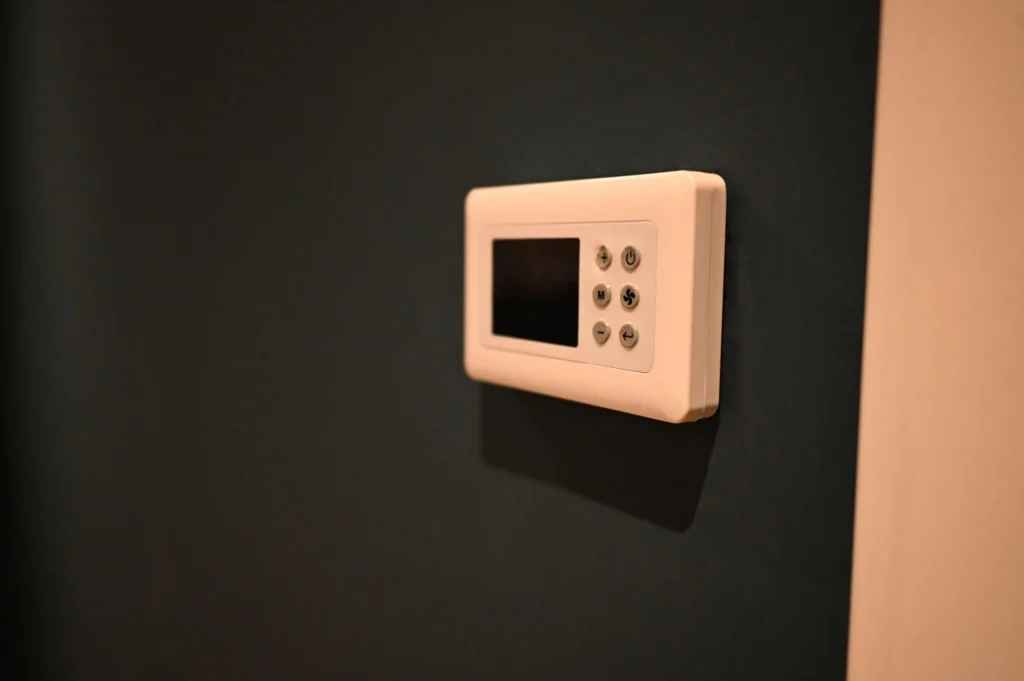 A white digital programmable home thermostat with a small black display screen and round control buttons mounted on a dark charcoal gray interior wall.